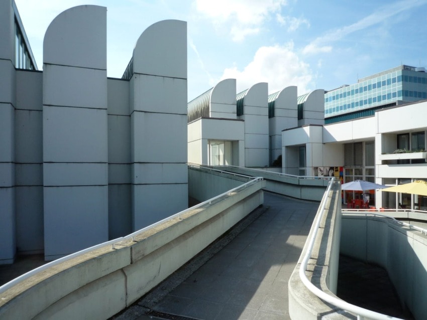 Modern architecture of a building complex with curved roof elements and a pedestrian walkway under a clear blue sky.