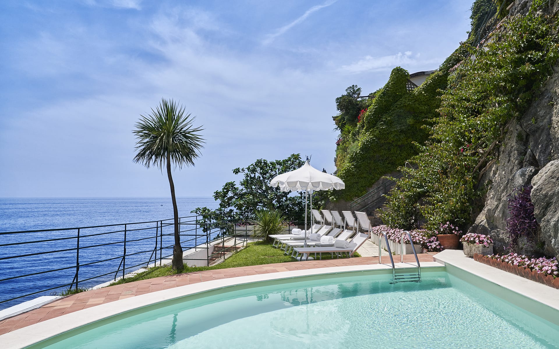 Infinity pool overlooking ocean with sun loungers and a palm tree, surrounded by lush greenery and a rocky hillside.