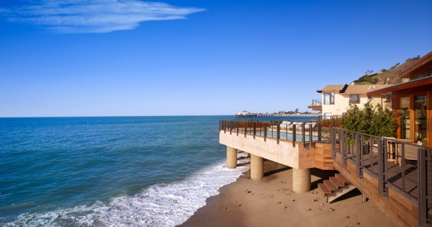 Ocean view from a wooden deck with clear blue skies and waves gently hitting the sandy shore in a coastal setting.