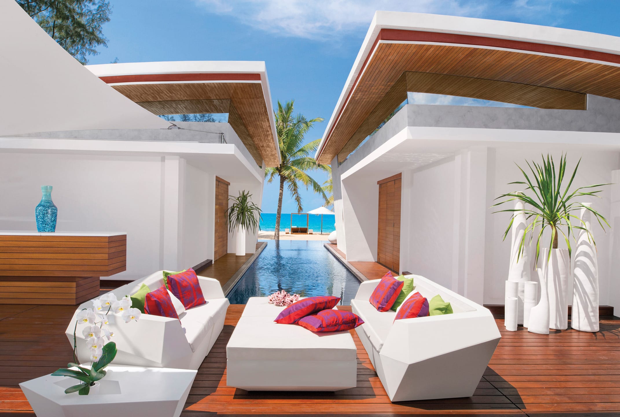 Modern beachfront villa with open-air patio, white furniture, vibrant cushions, pool, and ocean view.