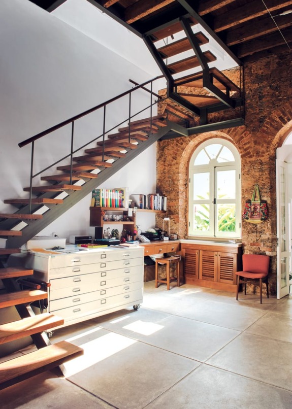 Loft space with wooden staircase, brick walls, an arched window, white cabinets, desk, and chair in a modern setting.