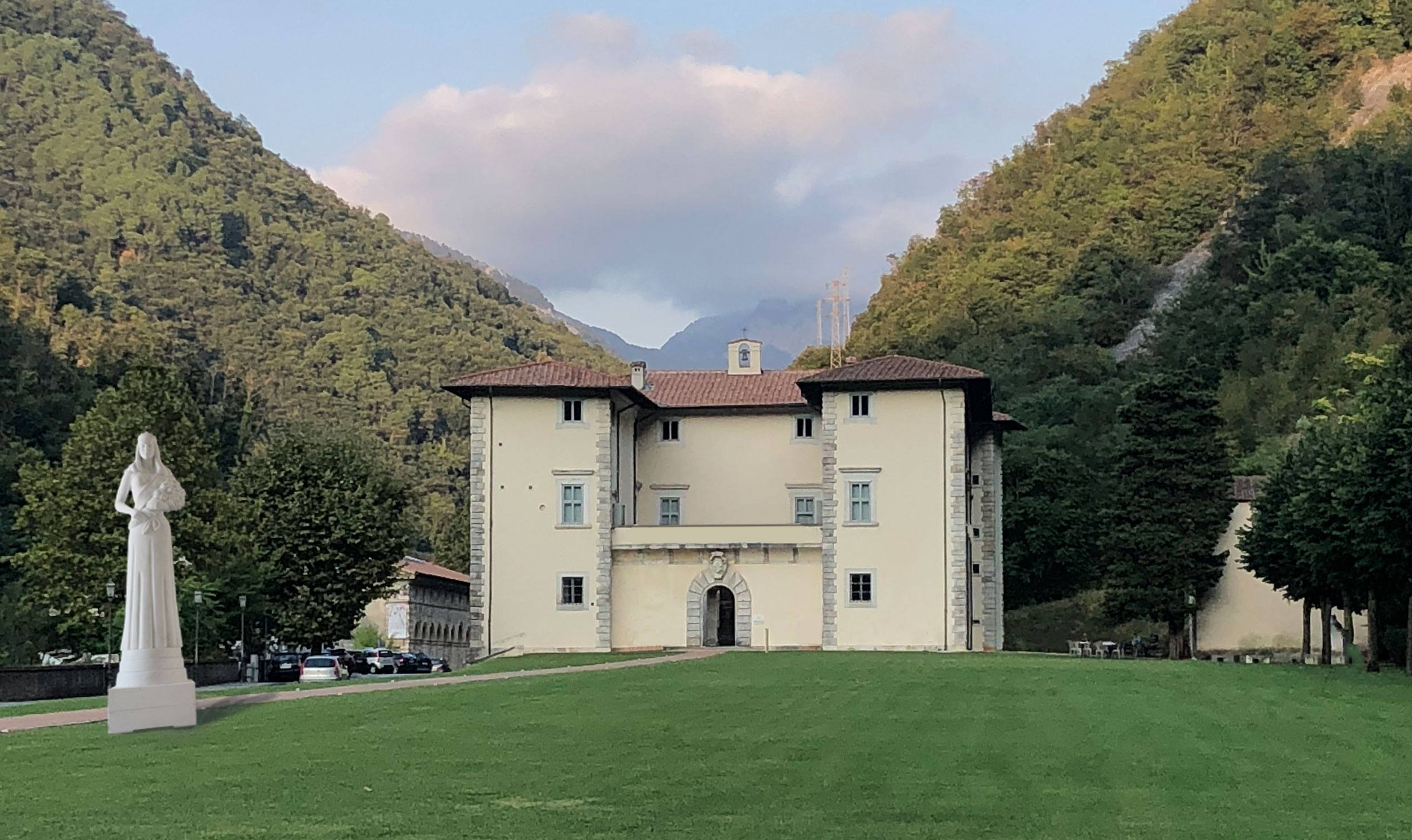 Historic villa surrounded by green hills with a statue in front, under a partly cloudy sky.
