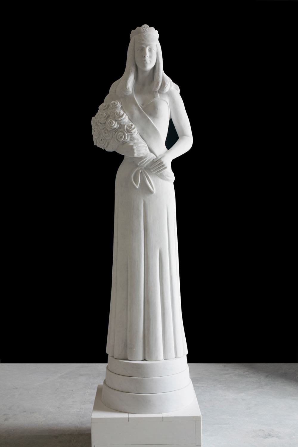 Sculpture of a woman in a gown holding a bouquet, standing on a pedestal against a black background.