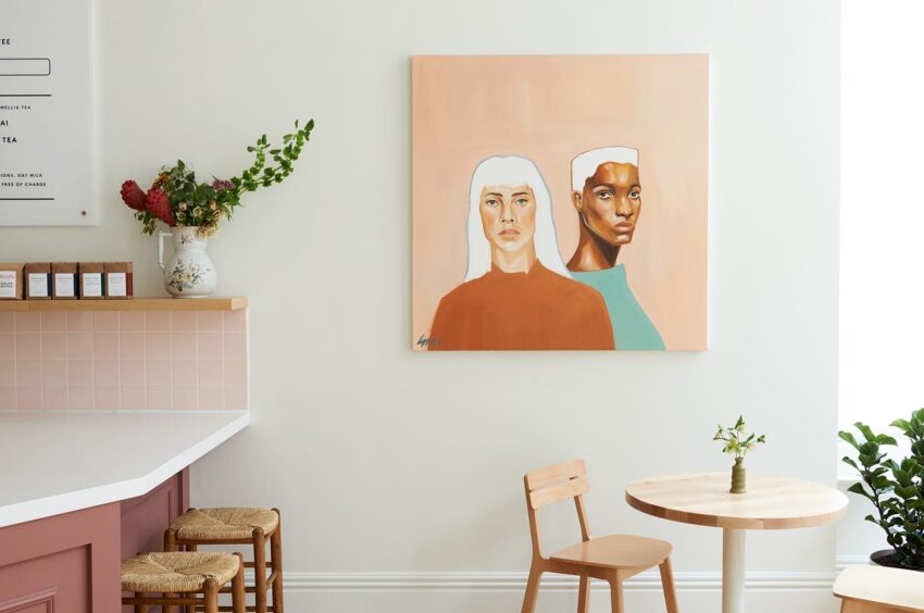 Modern cafe interior with a colorful portrait painting on the wall, featuring two people, and minimalist furniture arrangement.
