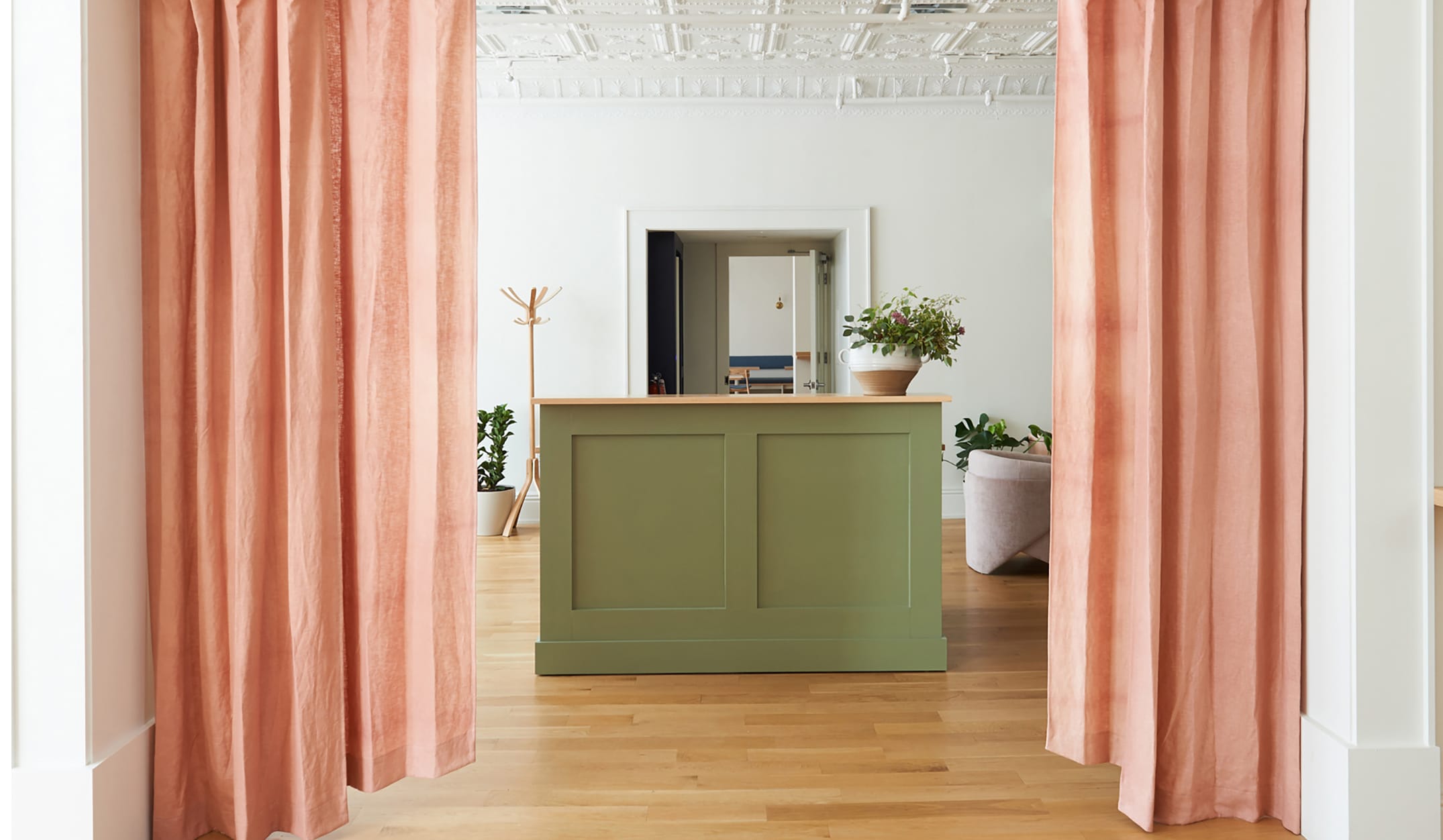 Green reception desk with potted plant, flanked by pink curtains in a brightly lit room with wooden floors and ceiling details.