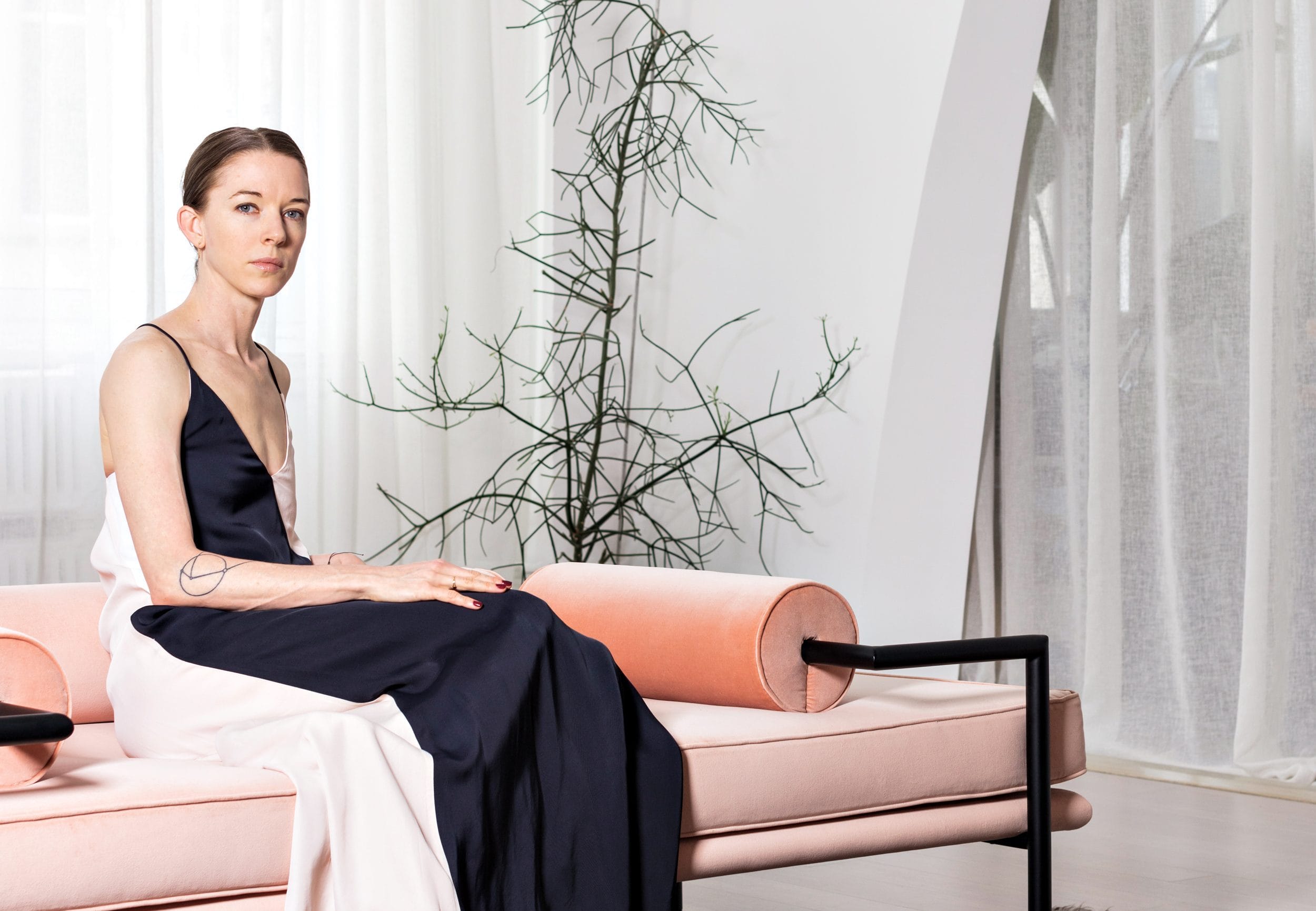 Woman in elegant dress sitting on a pink sofa in a modern, minimalistic room with a decorative plant.