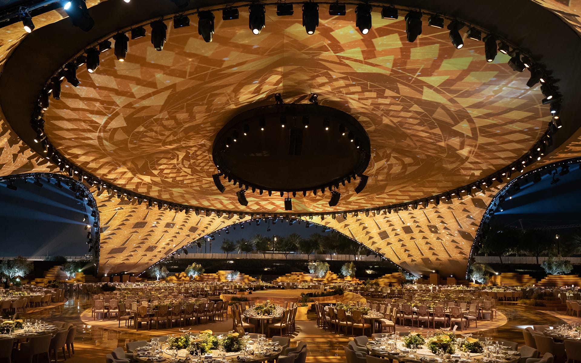 Spacious event venue with intricate ceiling pattern, elegant table arrangements, and ambient lighting for a large gathering.