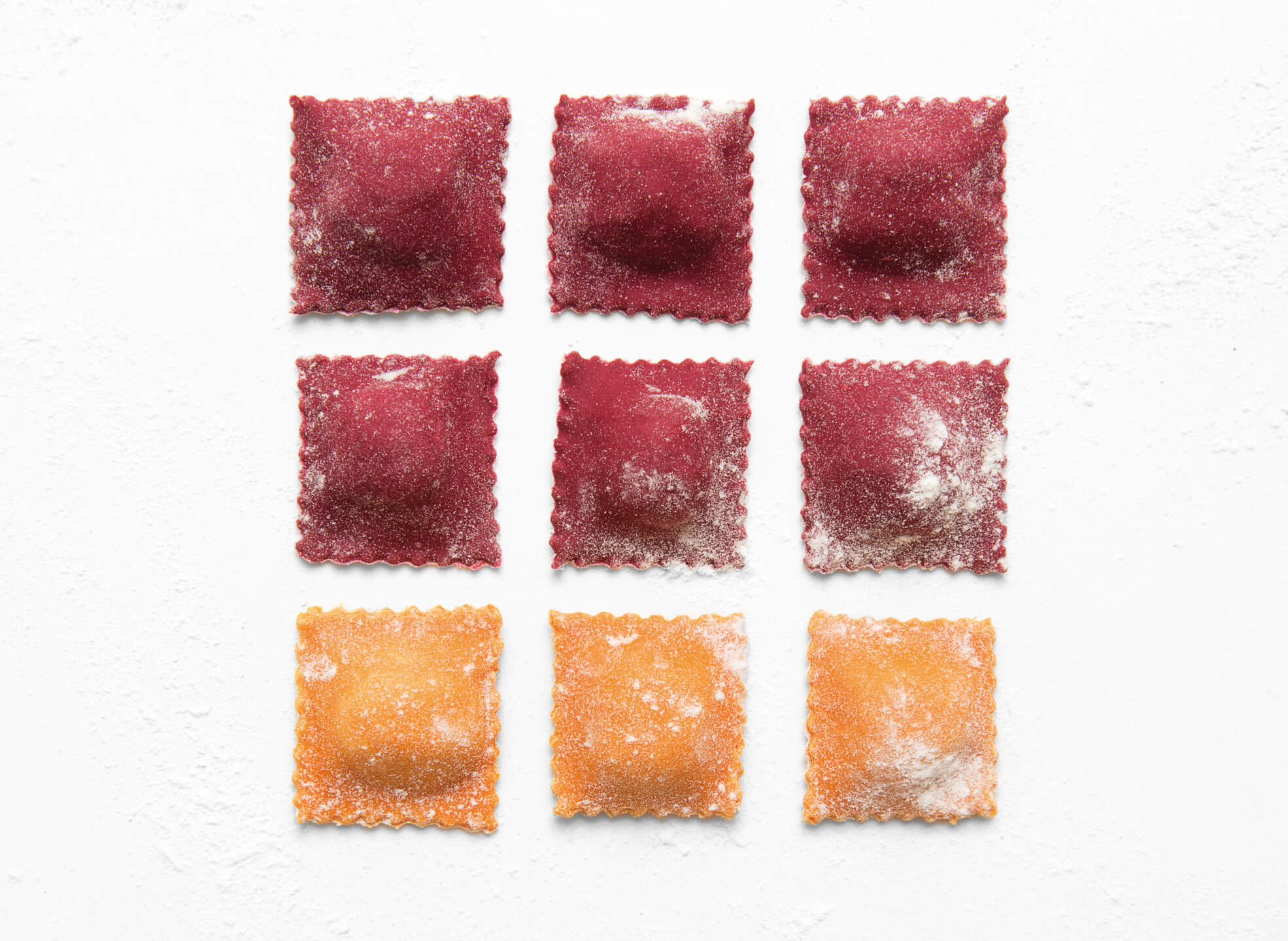 Nine colorful ravioli squares, arranged in a grid on a white surface, dusted with flour.