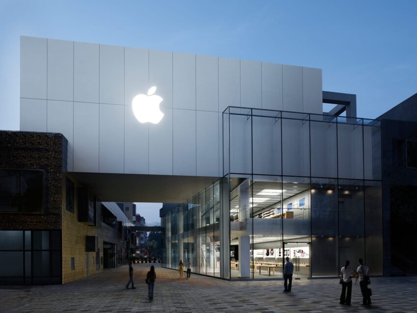 Modern glass building with a large illuminated apple logo on a white panel, people walking nearby at dusk.
