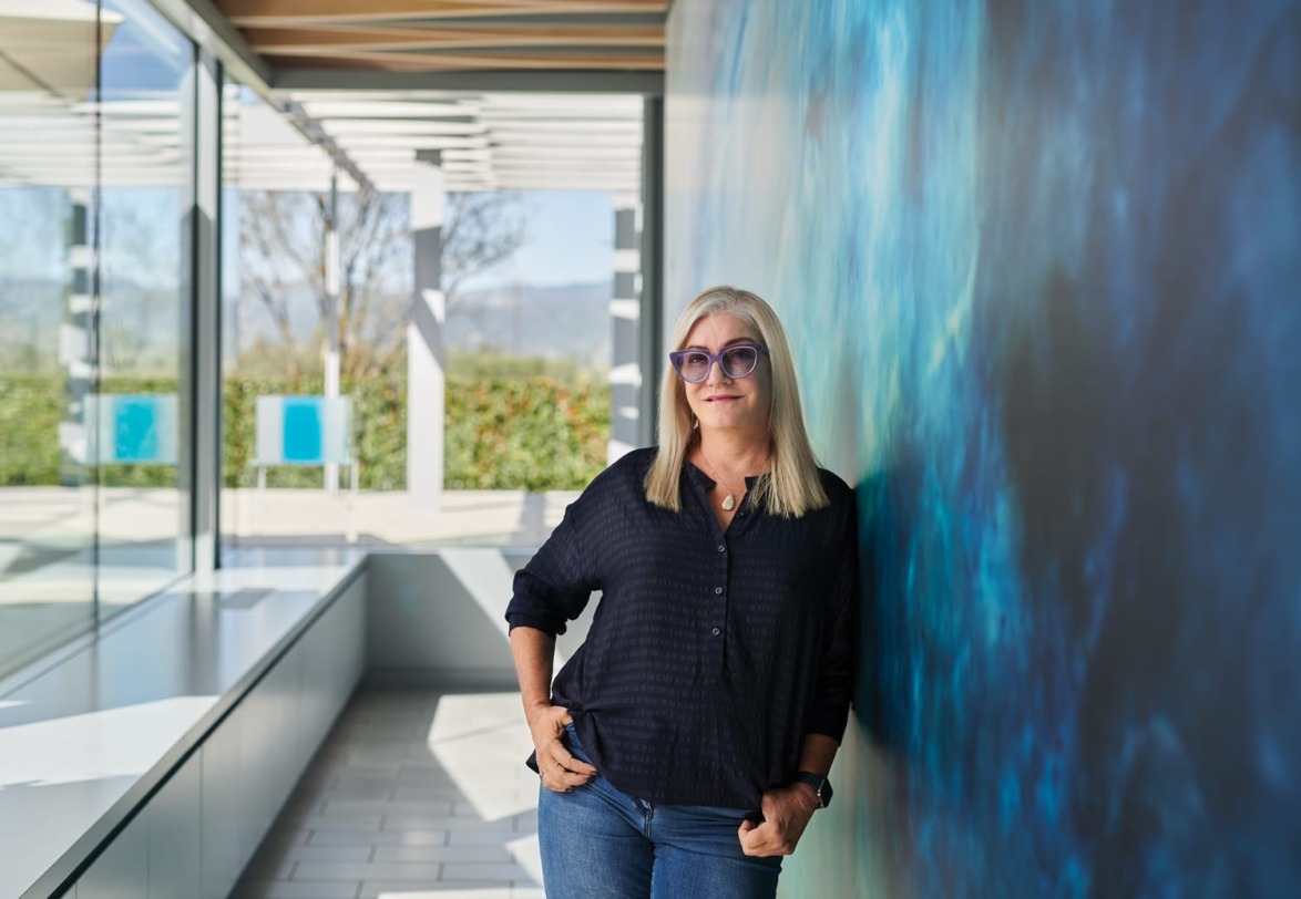 Woman with long gray hair and sunglasses stands by a blue wall in a modern building with large windows and natural light.