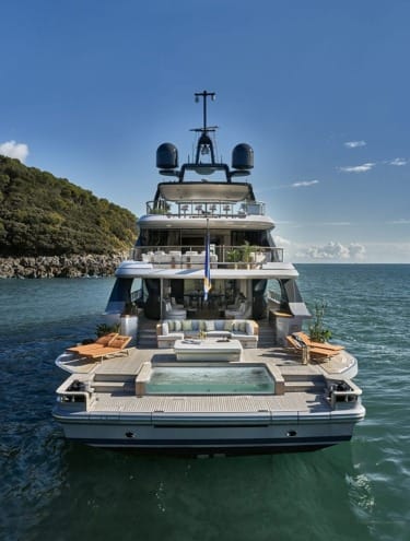 Luxury yacht with spacious deck and hot tub, cruising on open sea with clear blue sky and scenic coastline in the background.