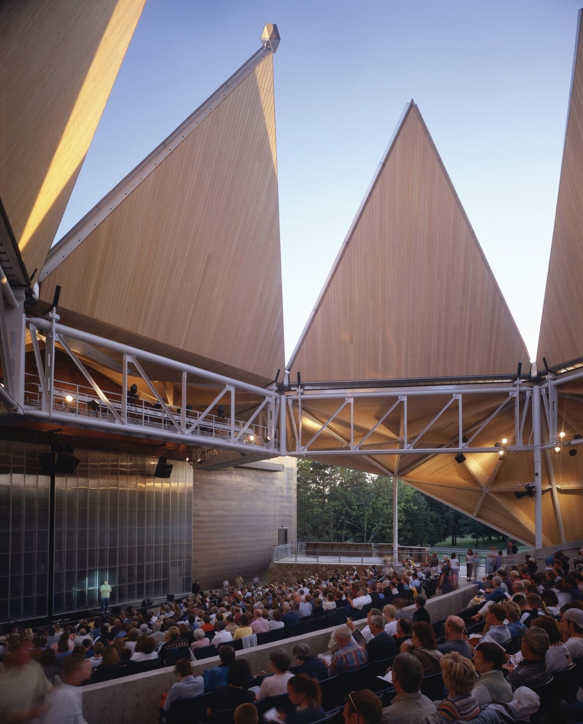 Outdoor concert venue with wooden triangular roofs and a seated audience enjoying an event at dusk.