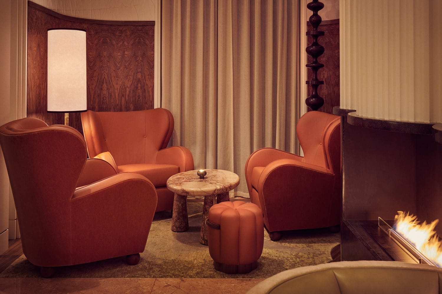 Cozy seating area with orange armchairs, round table, and fireplace, set against wood-paneled walls and curtains.