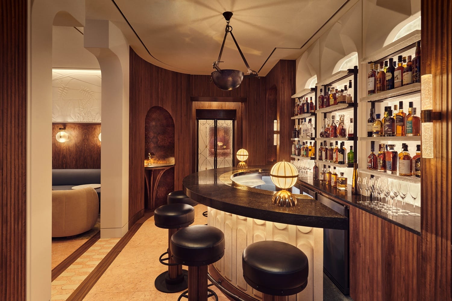 Cozy bar interior with a curved black countertop, upholstered stools, and shelves displaying various liquor bottles.