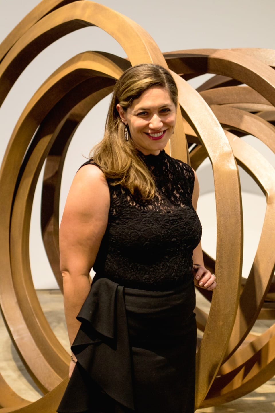 Person smiling, standing in front of a large abstract metal sculpture, wearing a black dress in an art gallery setting.