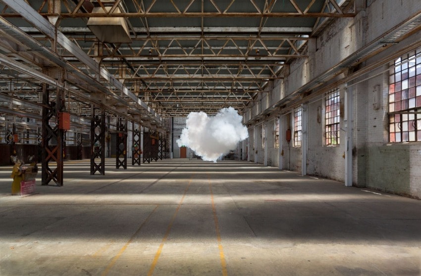 A large fluffy cloud floating inside an empty industrial warehouse with high ceilings and natural light streaming through windows.
