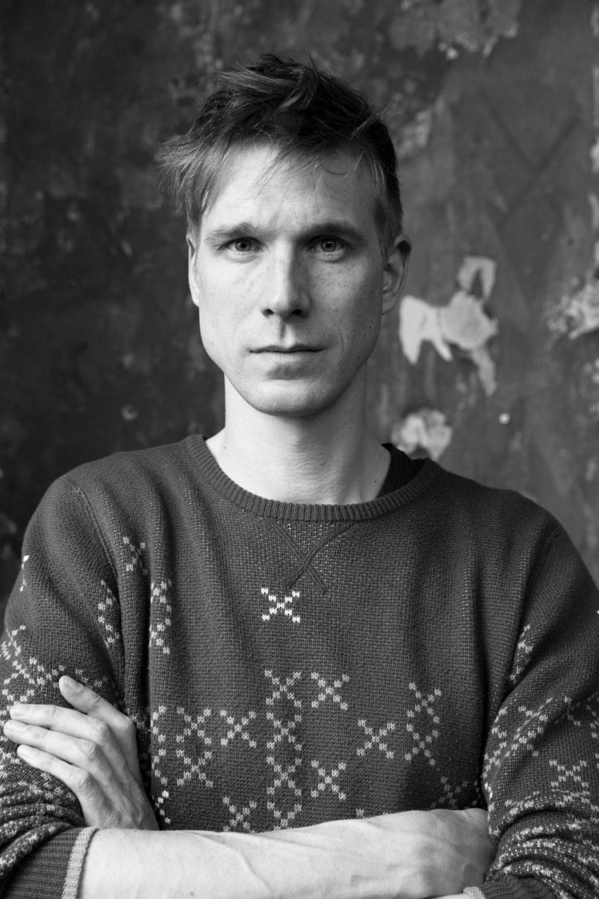 A person with short hair wearing a patterned sweater, standing with folded arms against a textured wall.