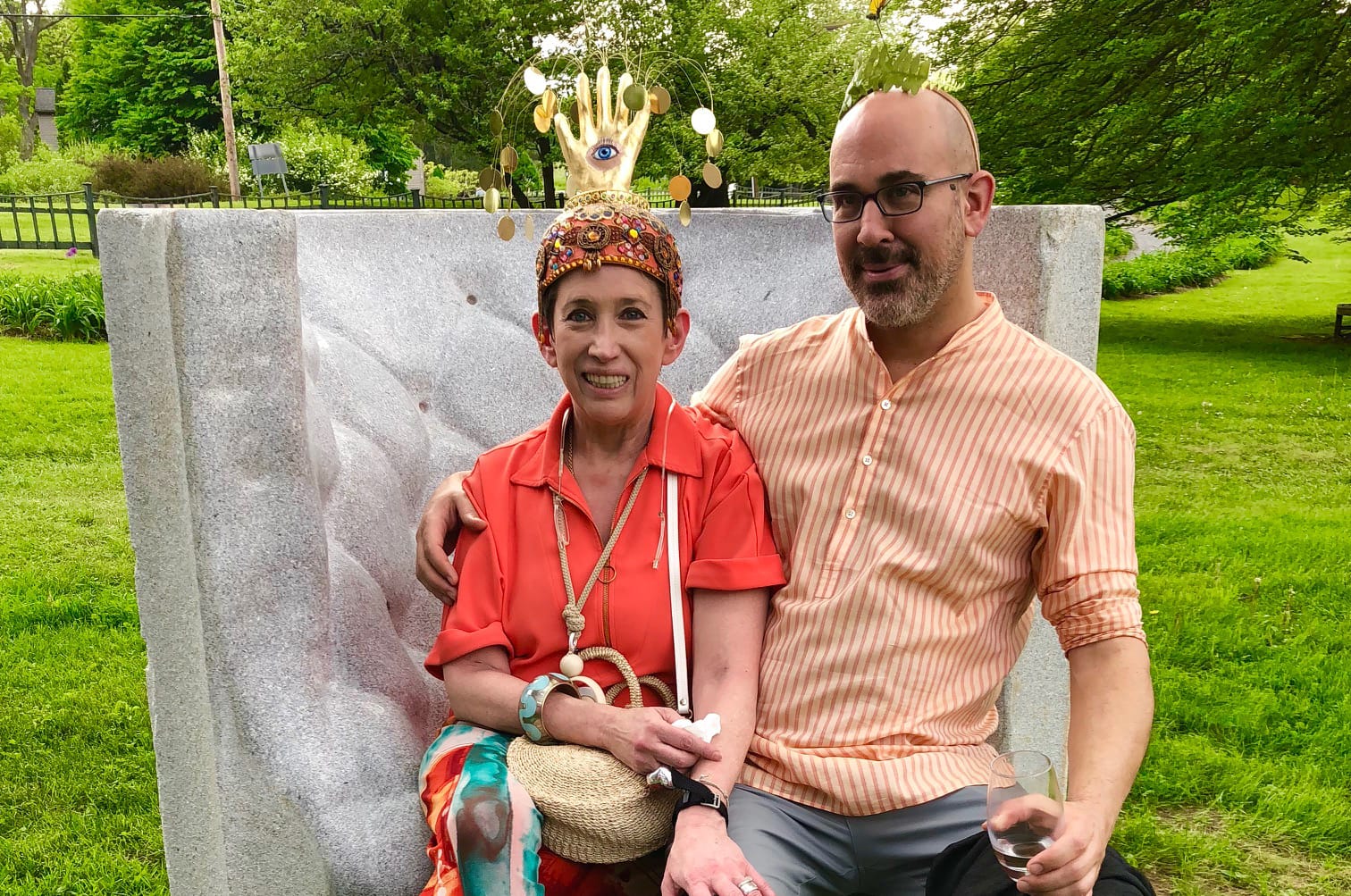 A woman in a colorful outfit and hat with a man sitting on a bench outdoors in a park setting.