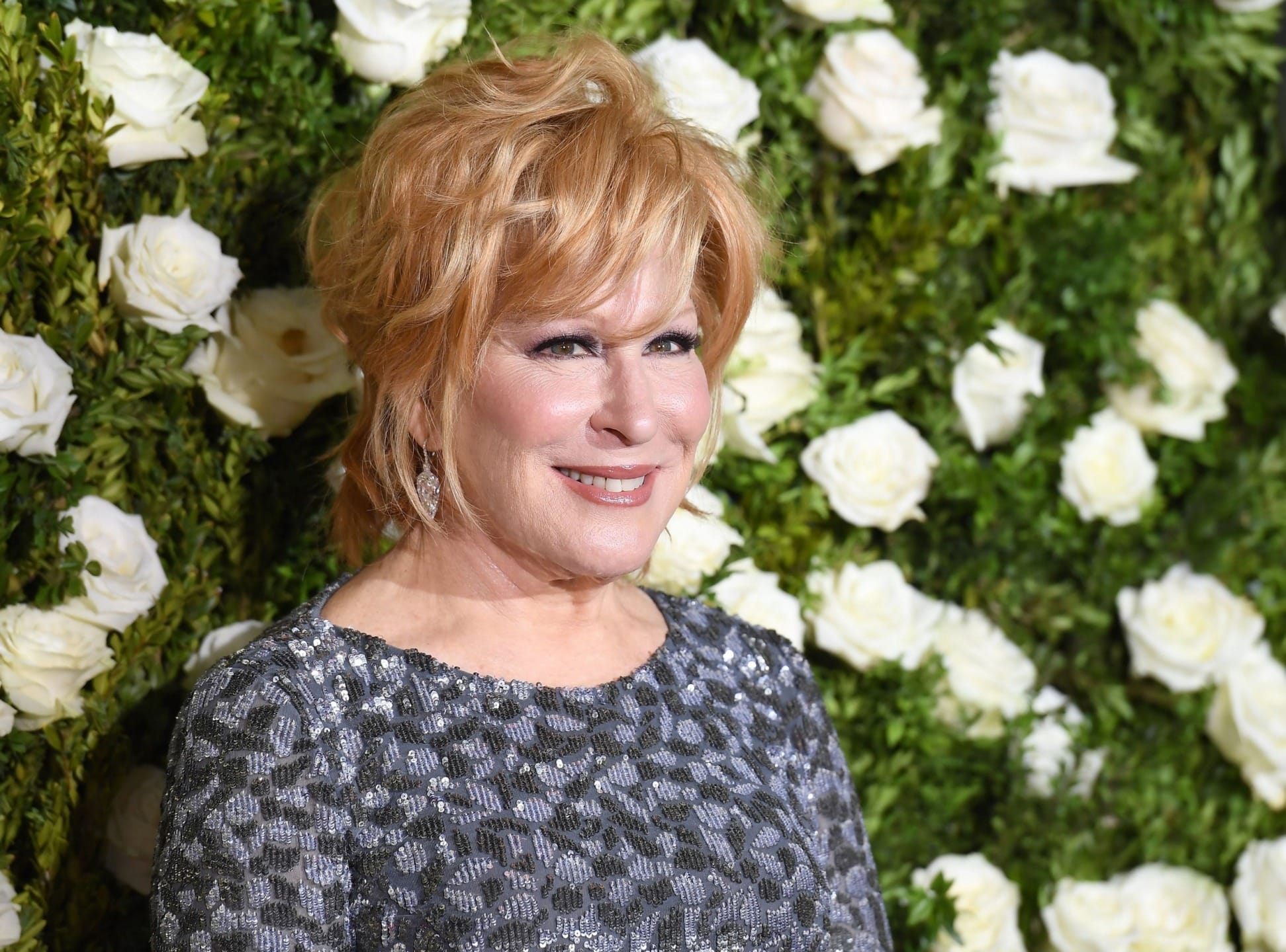 Person in a sparkly dress smiling in front of a wall of white roses.