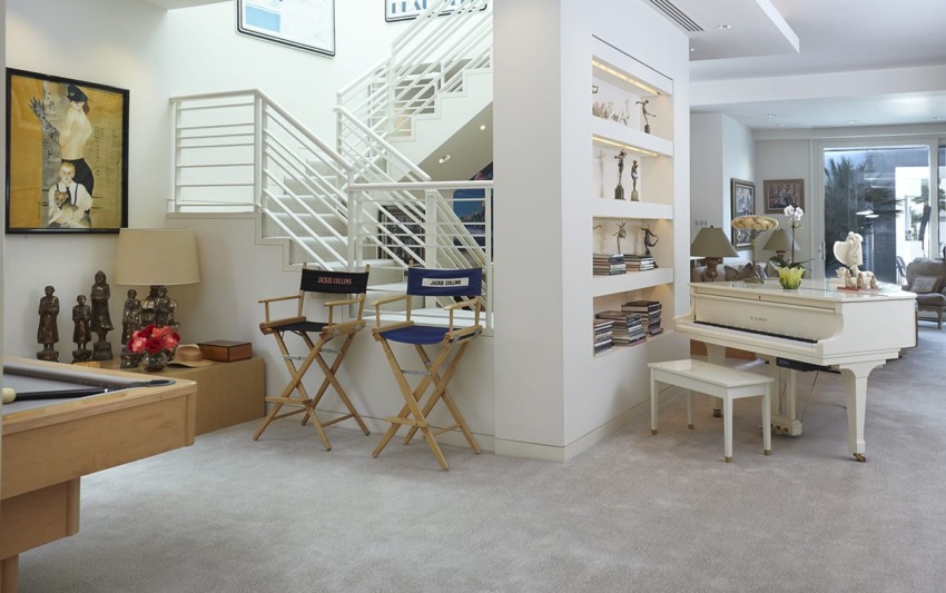 Modern living room with a white grand piano, director's chairs, decorative sculptures, and a staircase leading to an upper floor.