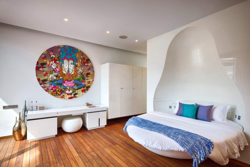 Modern bedroom with circular bed, colorful wall art, white furniture, and wooden floor.