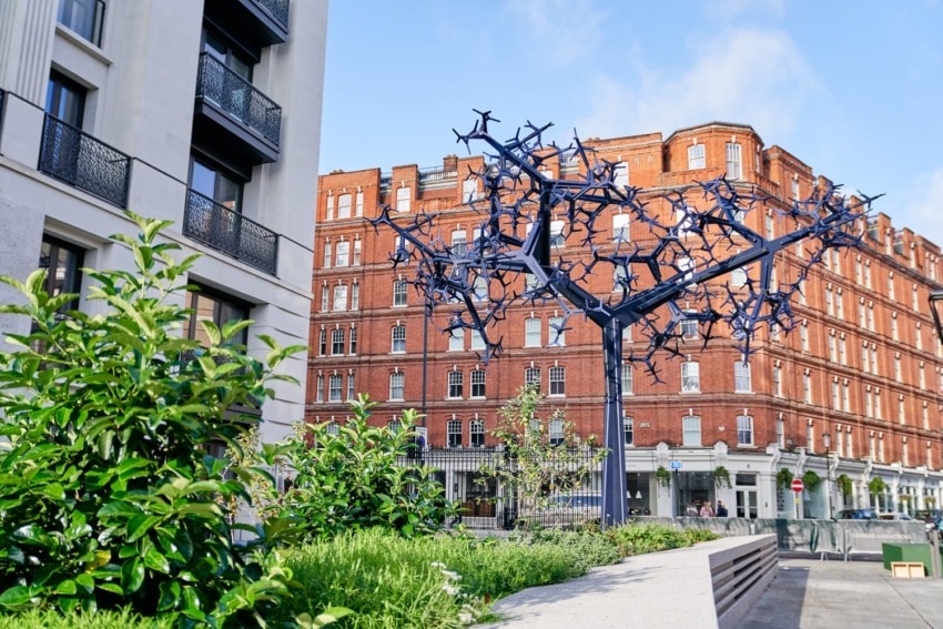 Modern metal sculpture resembling a tree in urban setting with red brick buildings and greenery surrounding.