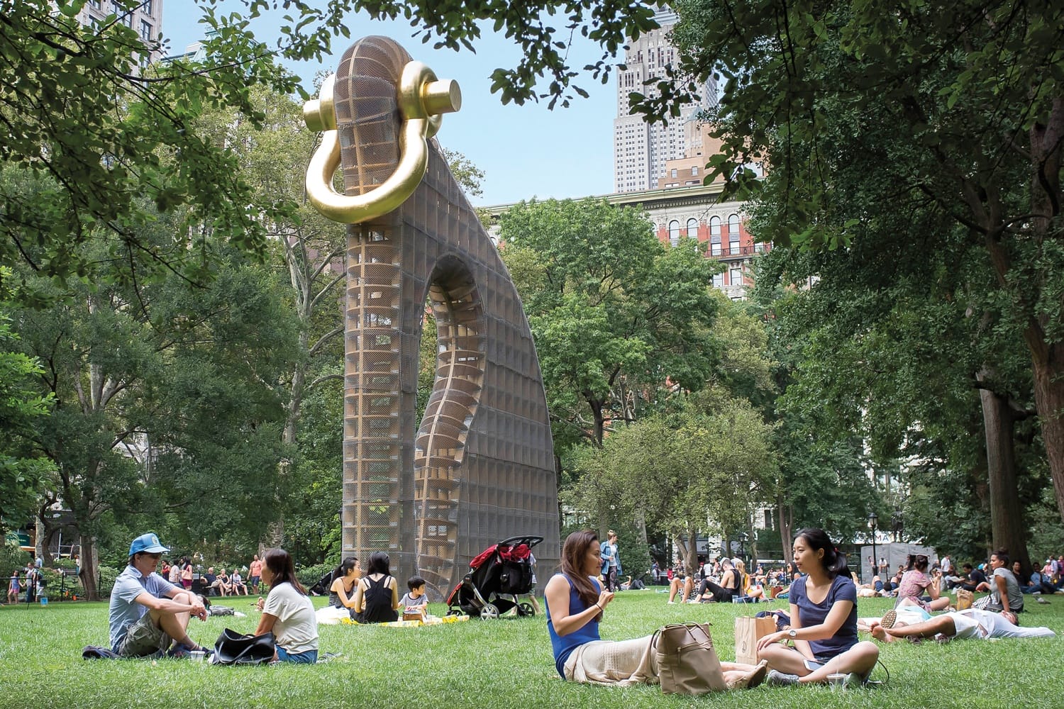 People relax on grass in a park near a large sculpture with a giant golden hoop, surrounded by trees and city buildings.