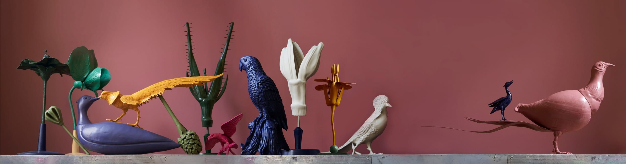 Colorful bird sculptures arranged on a shelf against a muted pink background with various plants and abstract shapes.