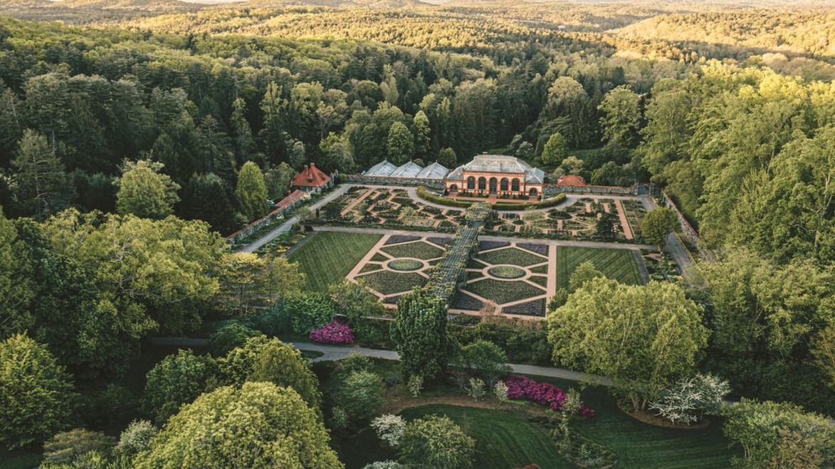 Aerial view of a large botanical garden with manicured lawns, colorful flower beds, and surrounding lush green forests.