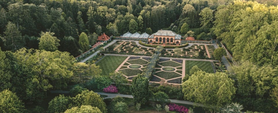 Aerial view of a large botanical garden with manicured lawns, colorful flower beds, and surrounding lush green forests.