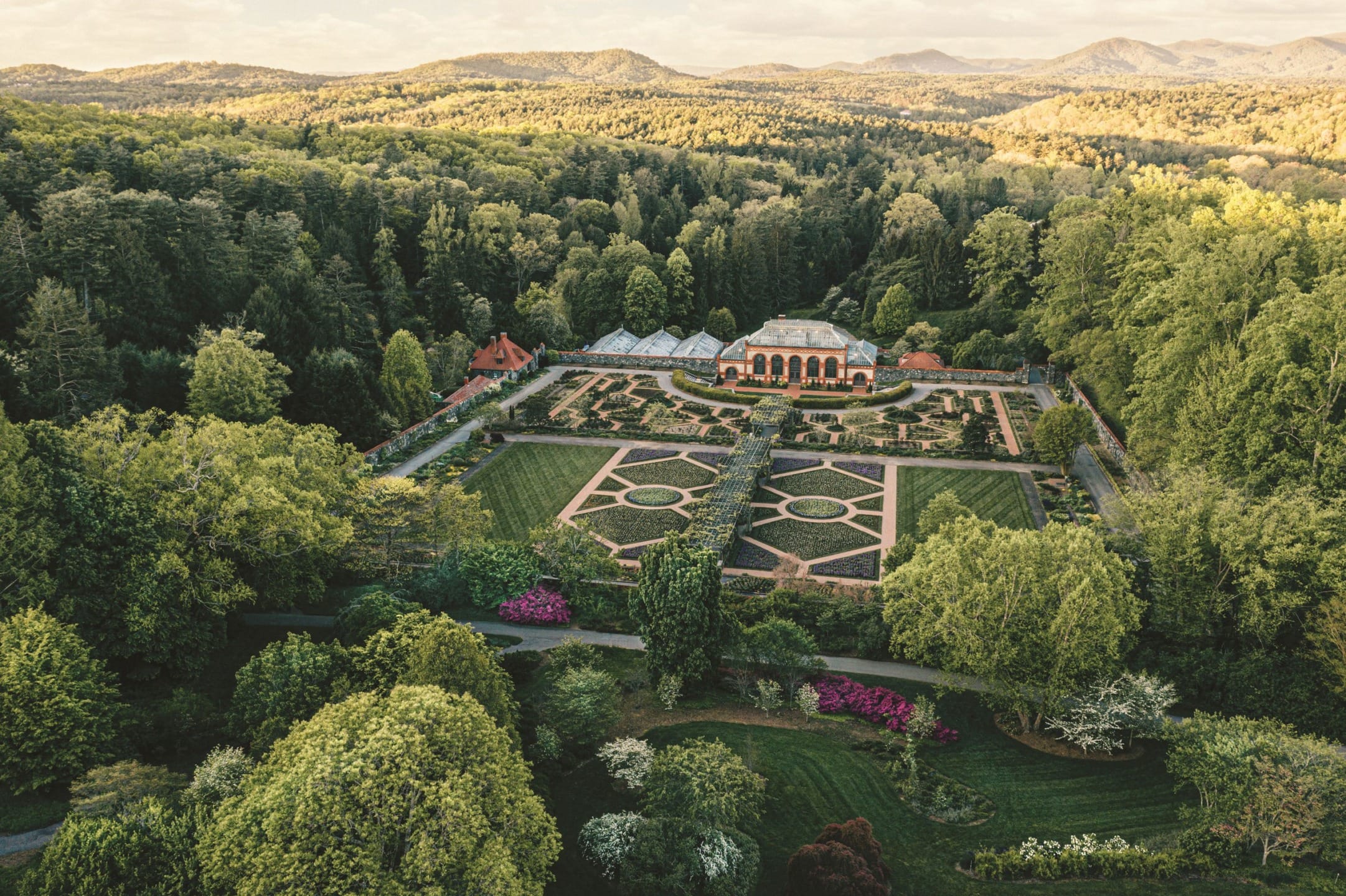Aerial view of a large botanical garden with manicured lawns, colorful flower beds, and surrounding lush green forests.