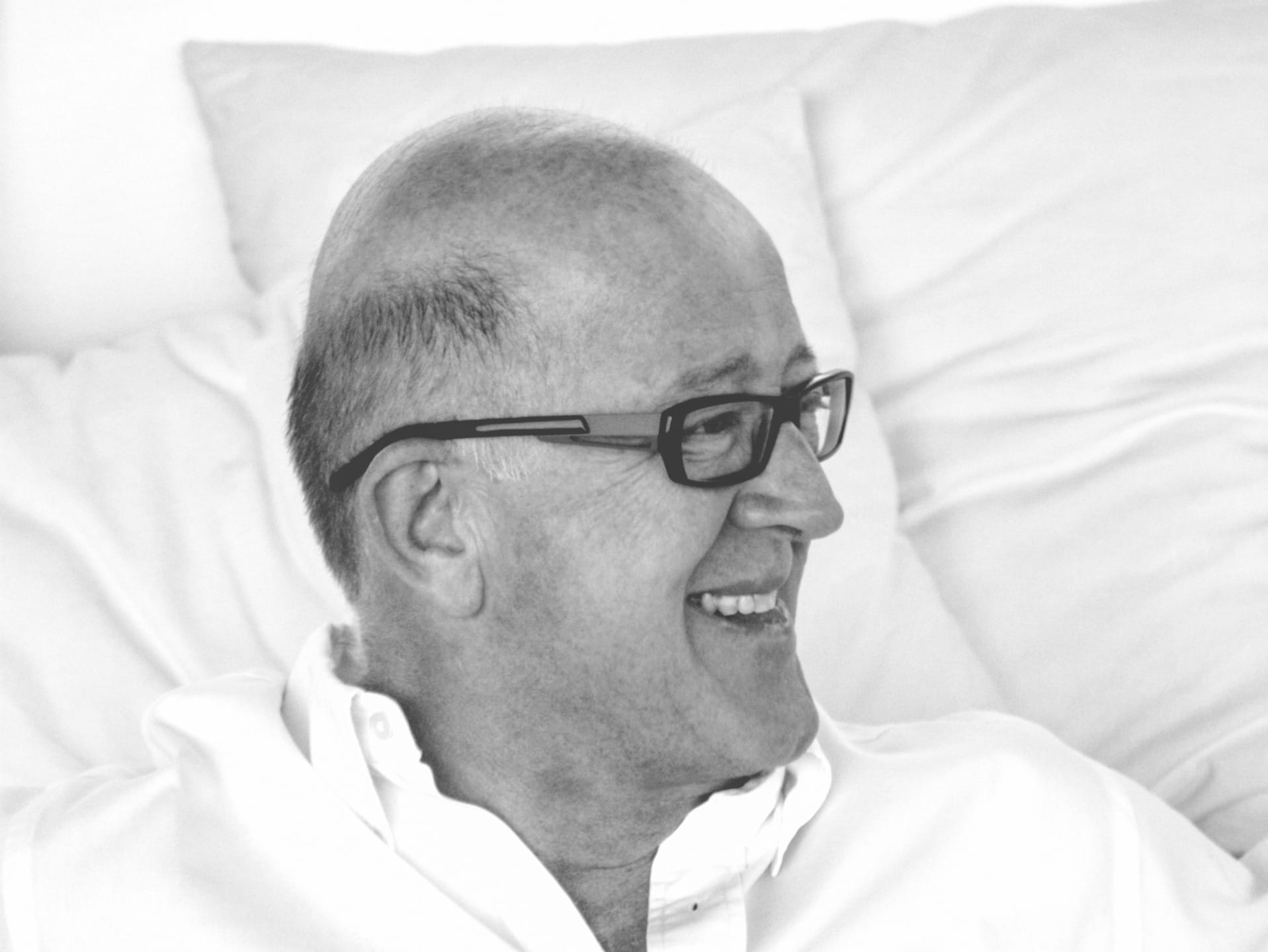 Man with glasses smiling and looking to the side, wearing a white shirt, in a black and white photo with a soft background.
