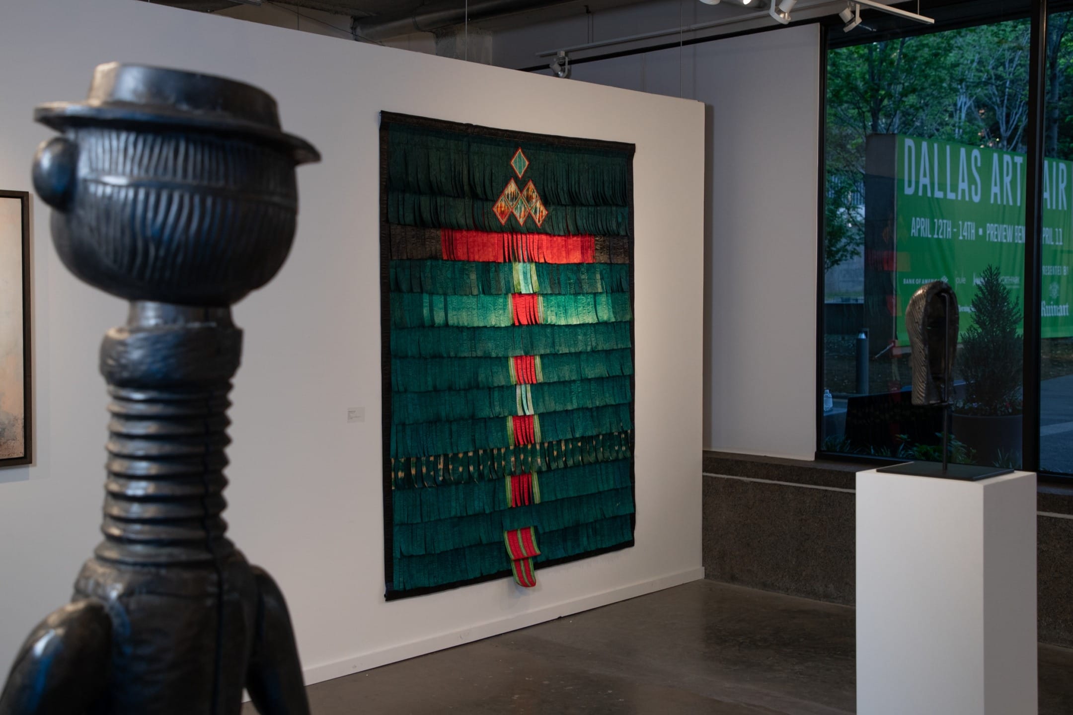 Sculpture in foreground with green textile art displayed on a gallery wall, large window showing outdoor sign in background.