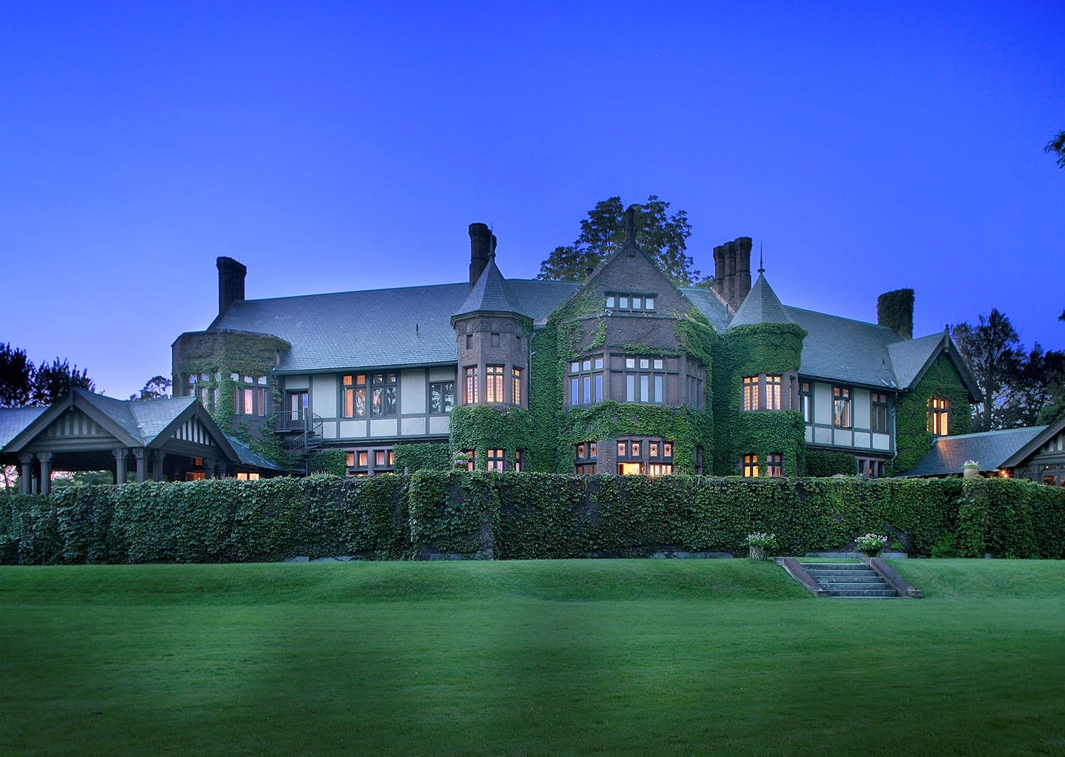 Large ivy-covered mansion with numerous windows, set against a twilight sky, surrounded by a manicured lawn.