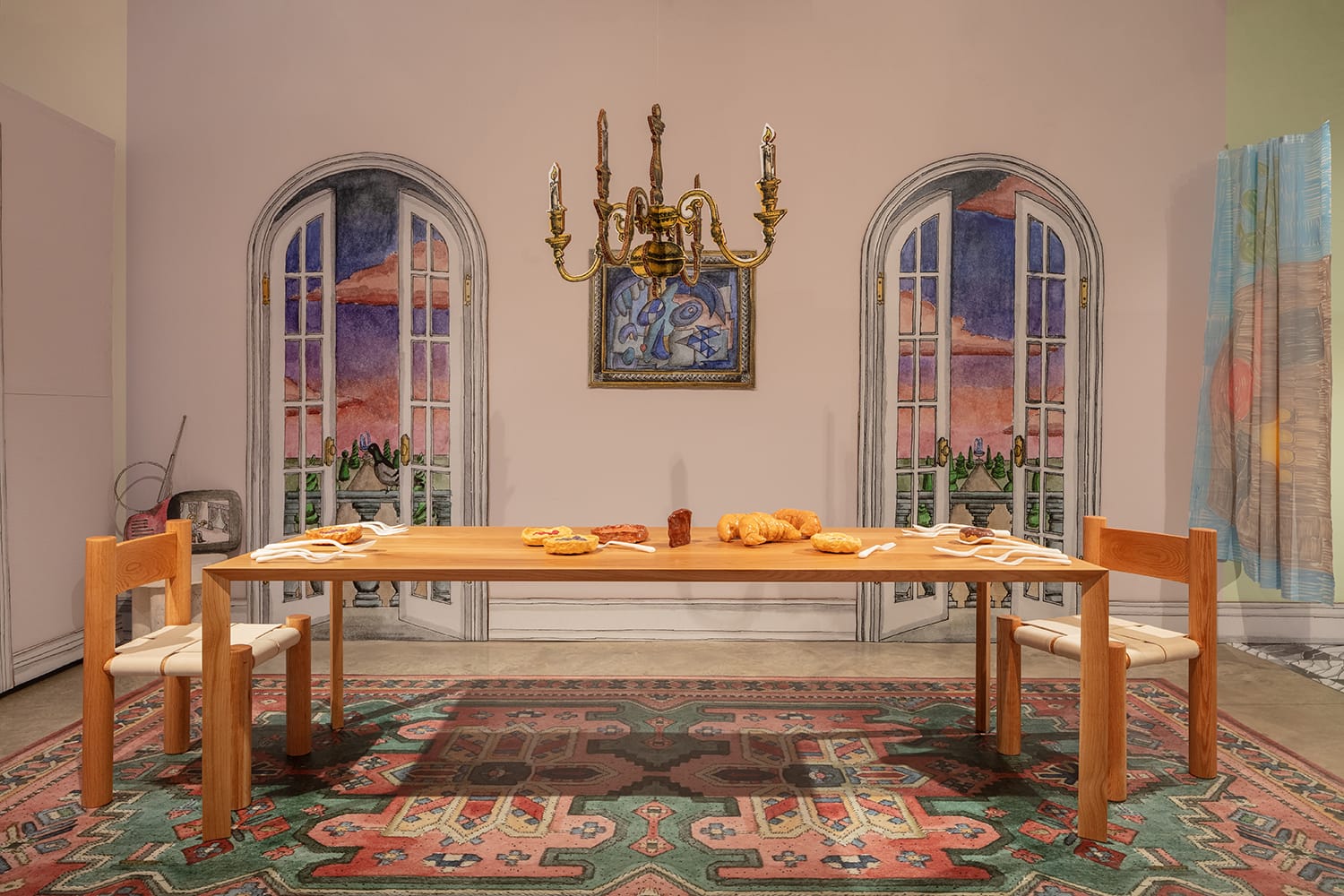 Dining table with bread and snacks, surrounded by chairs, under a chandelier, in front of painted doors and a colorful rug.