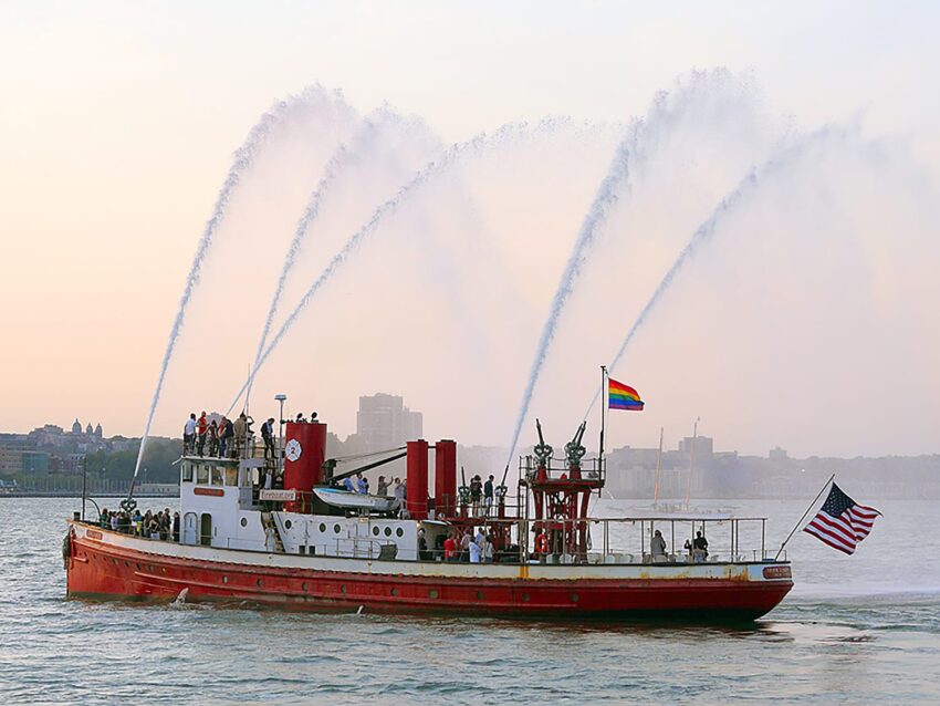 A boat on the water with multiple water jets spraying into the air, flying American and rainbow flags.