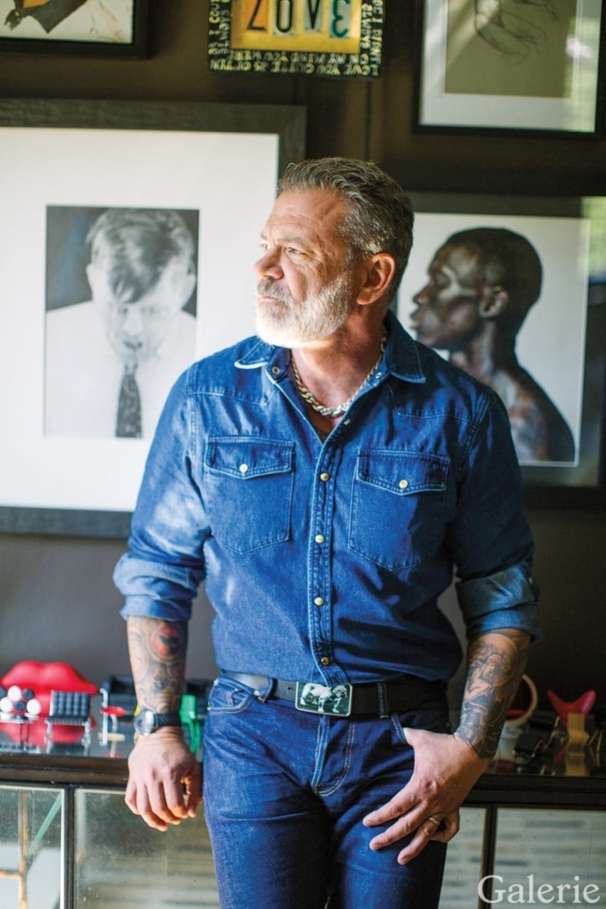 Man in denim outfit standing in art-filled room with tattooed arms, looking to the side.