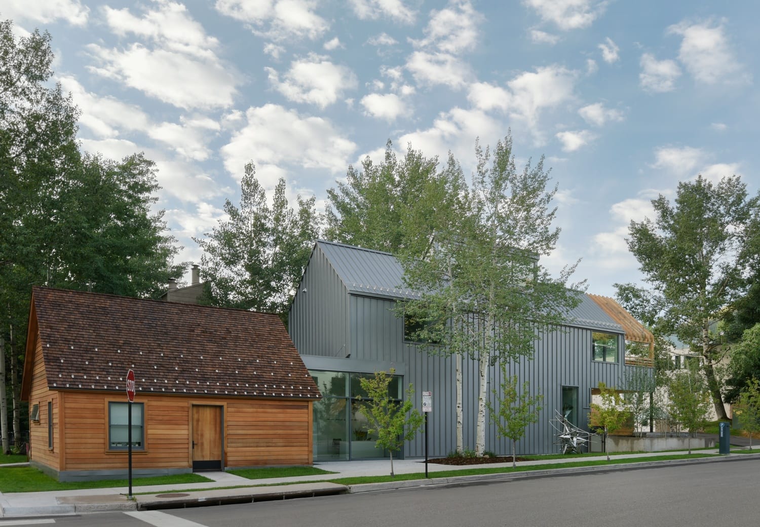 Modern house with metal exterior and wooden cabin surrounded by trees under a cloudy sky.
