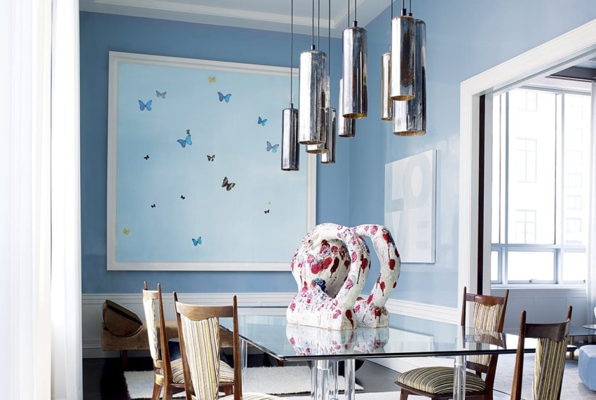Modern dining room with glass table, wooden chairs, abstract art, and pendant lights, featuring blue walls and large windows.