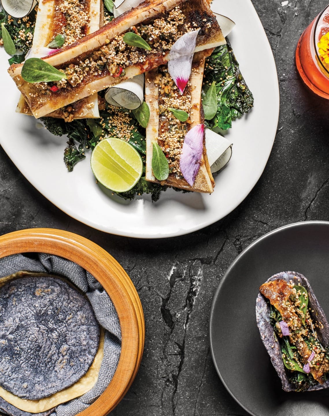 Gourmet dish with seasoned bone marrow, fresh greens, lime, and tortillas on a dark stone table setting.