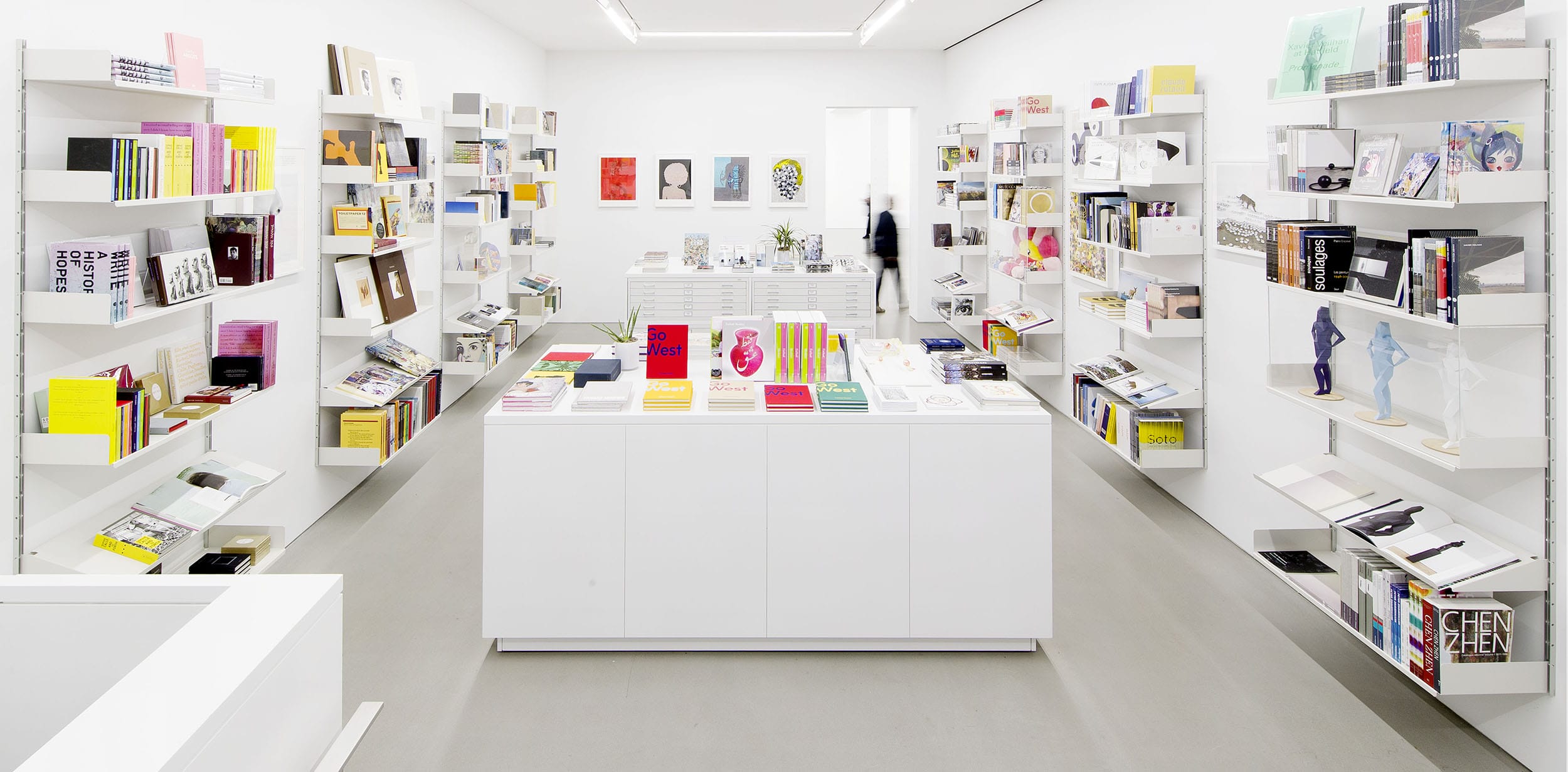 Modern bookstore with white shelves displaying colorful books and art pieces, centered around a white display table.
