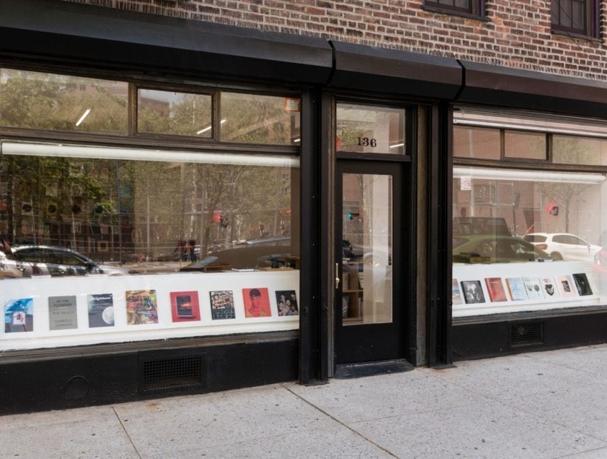 Storefront with large windows displaying books or magazines on shelves, set against a brick building and sidewalk scene.