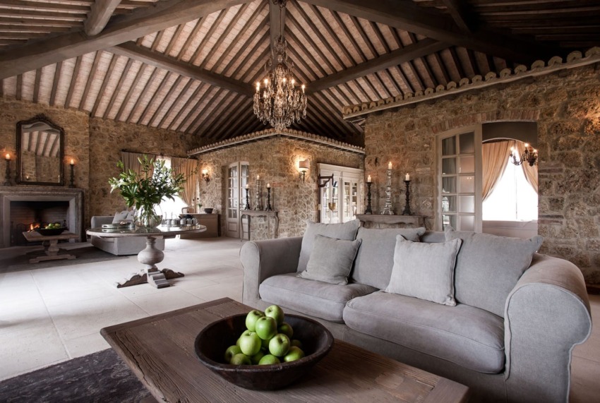 Rustic living room with stone walls, high wooden ceiling, sofa, chandelier, large table with flowers, and a bowl of green apples.