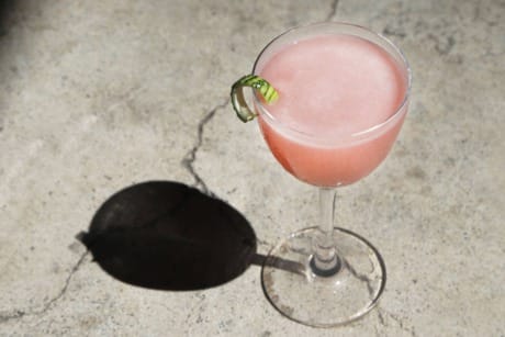 Pink cocktail in a glass with a cucumber garnish on a concrete surface.