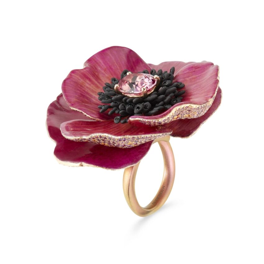 Gold ring featuring a large red flower with dark center and pink gemstone, viewed from an angle against a plain background.