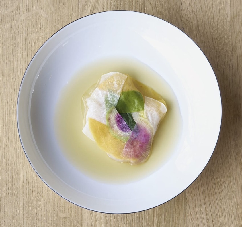 A gourmet dish featuring delicately arranged slices of colorful vegetables in a clear broth on a white plate.