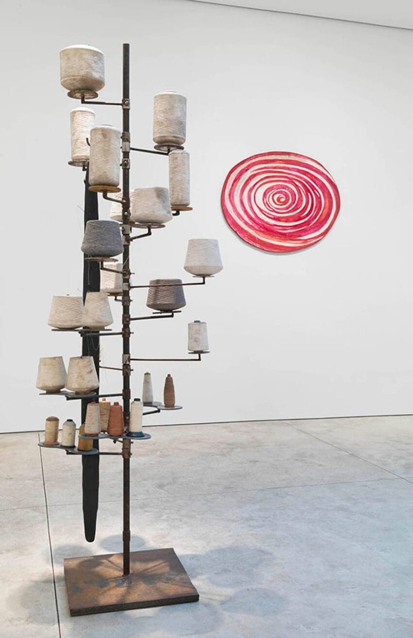 Abstract art installation with cylindrical objects on a metal structure and a red spiral painting on a white wall.