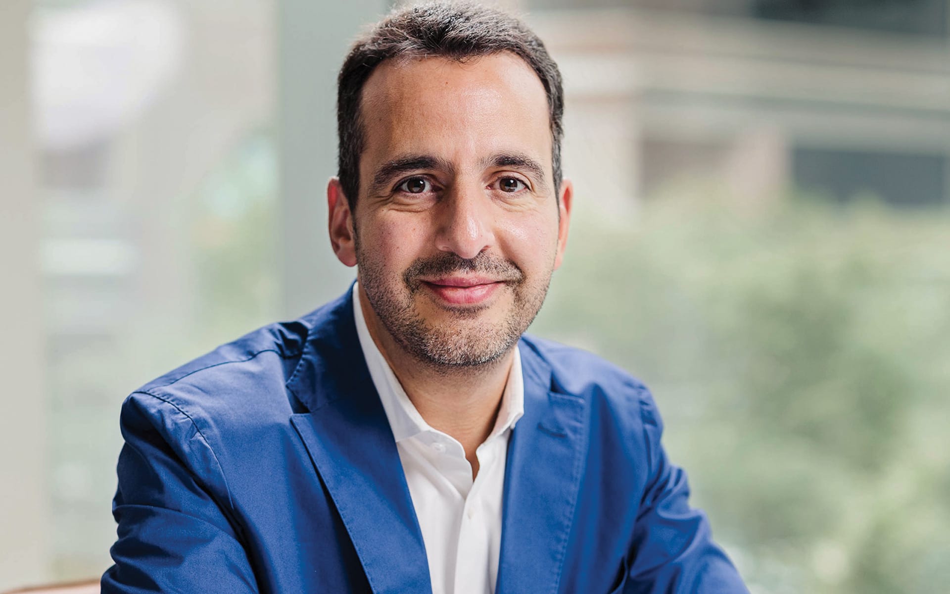Man in blue blazer and white shirt smiling with blurred background.