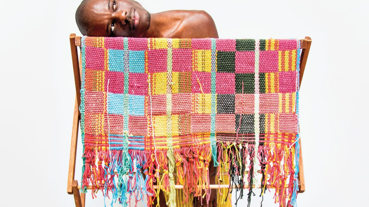 Man leaning on a wooden rack draped with a colorful, woven textile against a plain white background.
