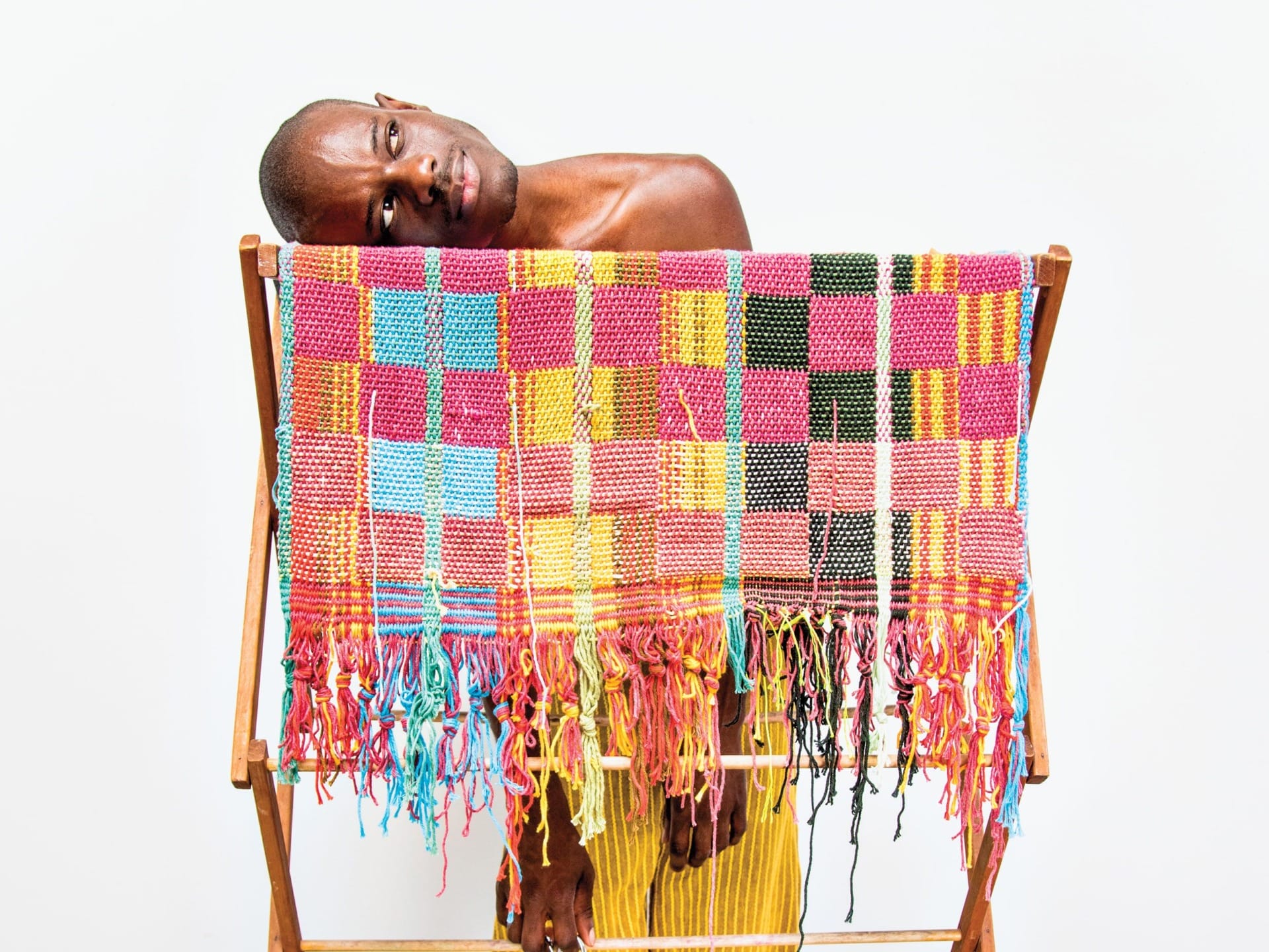 Man leaning on a wooden rack draped with a colorful, woven textile against a plain white background.